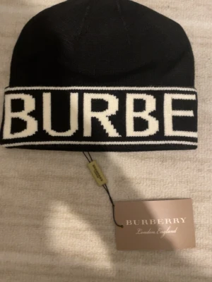 Svart mössa från Burberry - Snygg svart mössa från Burberry med vit logotyp i stora bokstäver runt hela uppviket. Klassisk och stilren design som passar till det mesta. Tillverkad i mjukt stickat material som håller dig varm under kalla dagar.