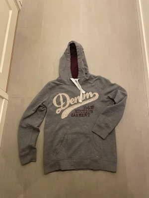 Grå hoodie med Denim-tryck - Snygg grå hoodie från Denim med stor vit och vinröd text på bröstet. Tillverkad i mjukt sweatshirtmaterial, perfekt för chill dagar. Tröjan står att den är i storlek L men är väldigt liten i storleken och passar snarare S eller XS! Priset är ej hugget i sten, skriv vid minsta fundering!!🤩🤩