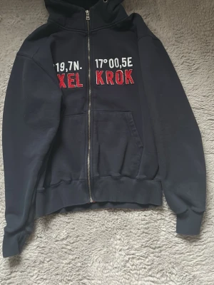 Mörkblå  hoodie med dragkedja - Mörkblå hoodie med dragkedja och huva. På bröstet står det 'BYXEL KROK' i stora röda bokstäver samt koordinater i vitt. Hoodien har känguruficka framtill och ribbade muddar. Perfekt för dig som gillar unika tryck och vill sticka ut. Själva modellen är lite ”sliten” i trycket men det ska vara så. Säljer då den ej kommer till användning 