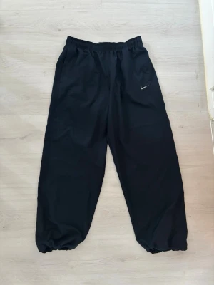 Svarta Nike mjukisbyxor med resår - Svarta mjukisbyxor från Nike med elastisk midja och logotyp på vänster sida. Byxorna har bred passform och resår vid benslut, vilket ger en avslappnad look. Perfekta för chill eller träning.