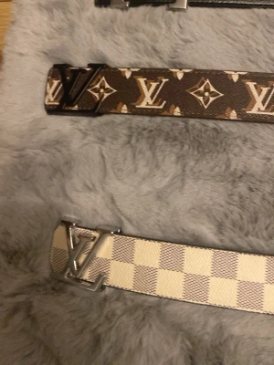 Louis Vuitton bälten med LV-spänne - Två Louis Vuitton bälten, ett i klassiskt brunt monogrammönster och ett i beige och vitt rutigt mönster. Båda har ett stort LV-spänne i silverfärg. Perfekta för att lyfta vilken outfit som helst.