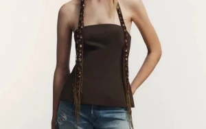 Brun korsettopp med dekorband - Snygg brun korsettopp med raka linjer och dekorativa band med paljetter och fransar som knyts i nacken. Toppen är axelbandslös och har en figurnära passform som framhäver siluetten. Perfekt för dig som vill sticka ut med coola detaljer.