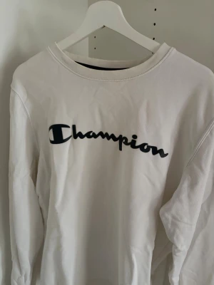 Vit sweatshirt från Champion - Säljer en vit sweatshirt från Champion med det klassiska logotrycket i svart på bröstet. Tröjan har rund halsringning och långa ärmar. Perfekt för en avslappnad och sportig stil.