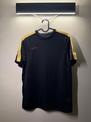 Svart Nike t-shirt med gula detaljer - Sportig svart t-shirt från Nike med gula paneler längs axlarna och ett rött Nike-märke på bröstet. Tillverkad i ett lätt och ventilerande material som passar perfekt till träning eller vardagsaktiviteter. Klassisk rund halsringning och korta ärmar.