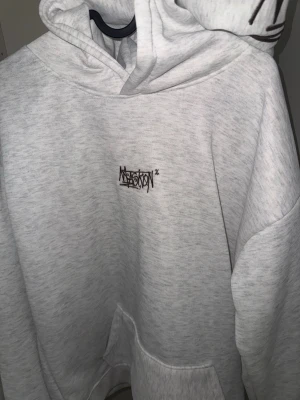 Ljusgrå hoodie med tryck - Säljer en ljusgrå hoodie från Nekstortion med svart tryck i graffiti-stil på bröstet och huvan. Hoodien har en klassisk känguruficka, ribbade muddar och är gjord i mjukt sweatshirtmaterial. Perfekt för en avslappnad streetwear-look.