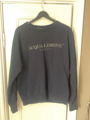 Mörkblå sweatshirt från Acqua Limone - Mörkblå sweatshirt från Acqua Limone med klassisk logga framtill och stor gul citron-grafik på ryggen. Tröjan har rund hals, ribbade muddar och är tillverkad i mjuk bomullsblandning. Perfekt för en avslappnad och trendig look.