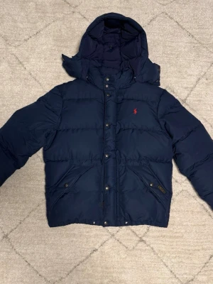 Mörkblå pufferjacka från Polo Ralph Lauren - Snygg mörkblå pufferjacka från Polo Ralph Lauren med huva och röd logga på bröstet. Jackan har tryckknappar, dragkedja och två snedställda fickor framtill. Perfekt för kalla vinterdagar och riktigt skön att bära.