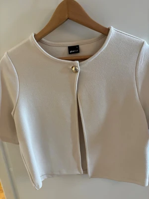 Kort beige kofta från Gina Tricot - Stilren kort kofta i beige från Gina Tricot med rund halsringning och en dekorativ guldfärgad knapp framtill. Koftan har korta ärmar och är gjord i ett mjukt, stretchigt material som ger en clean look.