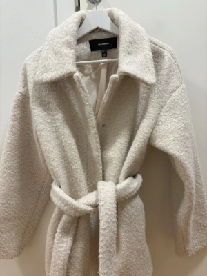Krämvit teddy-kappa från Vero Moda - Mysig krämvit teddy-kappa från Vero Moda med bred krage och knytskärp i midjan. Kappan har en fluffig textur, långa ärmar och är helfodrad för extra komfort. Perfekt för kyliga dagar när du vill ha en trendig och avslappnad look.