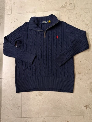 Mörkblå stickad tröja Polo Ralph Lauren - Mörkblå kabelstickad tröja från Polo Ralph Lauren med halv dragkedja och röd broderad logga på bröstet. Tröjan har ribbade muddar och hög krage, perfekt för en klassisk och stilren look. 