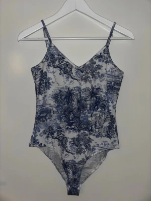 Vit och blå body med naturmönster - Snygg body med smala axelband och v-ringning, i vit bas med blått naturinspirerat mönster. Mjuk och stretchig känsla, perfekt att styla till jeans eller kjol. Ger en trendig och unik look med sitt detaljerade tryck.