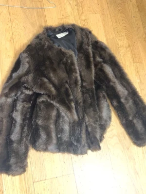 Mörkbrun pälsjacka i fuskpäls - Säljer en snygg mörkbrun pälsjacka i fuskpäls med klassisk rak passform och rundad krage. Jackan har långa ärmar och ett mjukt innerfoder som gör den extra skön under kalla dagar. Perfekt för dig som vill ha en trendig och varm look.