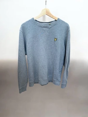 Ljusgrå sweatshirt från Lyle & Scott - Ljusgrå sweatshirt från Lyle & Scott med klassisk rund halsringning och det ikoniska gula fågelmärket broderat på bröstet. Tröjan har ribbade muddar vid ärmslut och nederkant, perfekt för en avslappnad och stilren look.