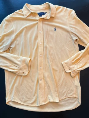 Ralph Lauren skjorta - Klassisk gul skjorta från Ralph Lauren med broderad blå logga på bröstet. Skjortan har lång ärm, knappar framtill och en mjuk krage. Tillverkad i ett tunt, bekvämt bomullstyg som känns skönt mot huden. Perfekt för en stilren och avslappnad look.