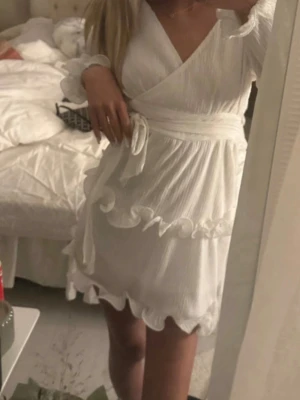 Vit omlottklänning med volanger - Supersöt vit omlottklänning med volangdetaljer och knyt i midjan. Klänningen har långärmade ärmar med volangkant och är tillverkad i ett lätt, strukturerat tyg som ger en luftig känsla. Perfekt för sommarens alla tillfällen!