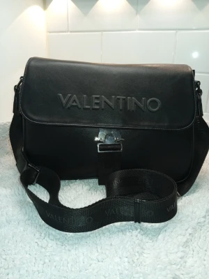 Svart axelväska från Valentino - Snygg svart axelväska från Valentino med präglad logga på framsidan och justerbar axelrem. Väskan har ett stort huvudfack, innerficka med dragkedja och fack för kreditkort och stängs med ett metallspänne. Tillverkad i slätt konstläder med textilfoder.