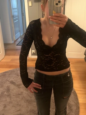 Svart spetsblus - Säljer en svart långärmad blus i spets med djup v-ringning och scallop-kant. Blusen är croppad och har ett transparent, elegant utseende som passar perfekt till jeans eller kjol. Perfekt för dig som gillar att sticka ut med detaljer och stil. Ett lite större hål i armhålan