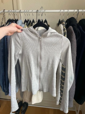 Ljusgrå ribbad hoodie med dragkedja - Säljer en ljusgrå hoodie i ribbat material med dragkedja framtill och huva. Hoodien har långa ärmar och en smal passform, perfekt för en avslappnad och stilren look. Passar till det mesta och är superenkel att matcha.