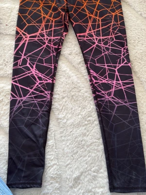 Svarta leggings med neonmönster - Snygga leggings med svart bas och grafiskt mönster i neonrosa och orange. Tight passform och stretchigt material som sitter skönt. Perfekta för dig som gillar färg och vill sticka ut. Mönstret ger en cool och modern vibe.