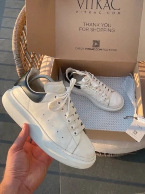 Vita Alexander McQueen sneakers - Säljer ett par vita sneakers från Alexander McQueen med chunky sula och svart detalj på hälen. Skorna har klassisk snörning, perforerade sidor och är tillverkade i läder. Perfekta för dig som gillar stilrena och trendiga skor.