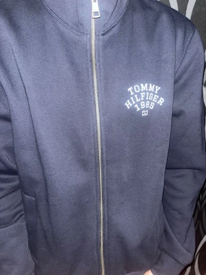 Mörkblå zip hoodie Tommy Hilfiger - Mörkblå hoodie från Tommy Hilfiger med dragkedja framtill och tryckt logga på bröstet där det står 'Tommy Hilfiger 1985'. Hoodien har långa ärmar och är tillverkad i mjukt sweatshirtmaterial. Perfekt för en avslappnad och stilren look.