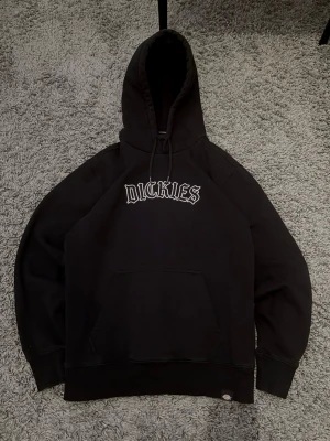Svart Dickies hoodie med tryck - Säljer en svart hoodie från Dickies med stor huva och dragsko. Framtill finns ett vitt Dickies-tryck i streetstil och en klassisk magficka. Hoodien är gjord i mjukt bomullsmaterial och har ribbade muddar vid ärmar och nederkant.