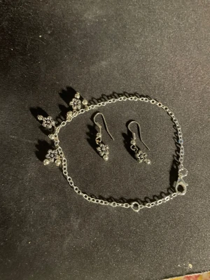 Silverfärgat smyckeset med blommor - Säljer ett set med armband och örhängen i silverfärgad metall. Båda delarna har små hängande blommor med detaljerade blad och pärlliknande inslag. Perfekt för dig som gillar söta och unika accessoarer med blommotiv. OBS! Materialet är okänt, kan ej garantera att det är rostfritt.