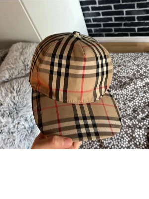 Beige keps med rutigt mönster - Säljer en klassisk beige keps från Burberry med det ikoniska rutiga mönstret i svart, vitt och rött. Kepsen har böjd skärm och justerbar rem bak. Perfekt accessoar för dig som vill ha en tidlös och trendig look.