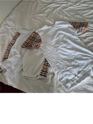 Vitt set med rutiga detaljer - Säljer ett vitt set med t-shirt och shorts. Båda plaggen har klassiska beige, svarta och röda rutiga detaljer på ärmar och sidor. Setet har en avslappnad passform och är perfekt för en trendig look. Materialet känns mjukt och bekvämt.