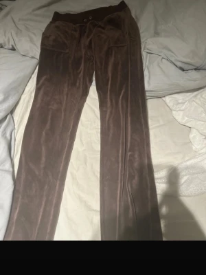 Bruna mjukisbyxor i velour - Säljer ett par bruna mjukisbyxor i mjuk velour med bred resår i midjan och två fickor med lock och knappar bak. Byxorna har raka ben och en avslappnad passform, perfekta för chill dagar. Passar dig som gillar en retro och bekväm stil.