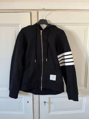 Thom Browne Zip Hoodie - Snygg svart hoodie från Thom Browne med dragkedja framtill, huva med dragsko och fyra vita ränder på ena ärmen. Tillverkad i mjuk bomull och har en liten vit patch med logga nedtill. Perfekt för en avslappnad streetwear-look. Storlek M men sitter som S