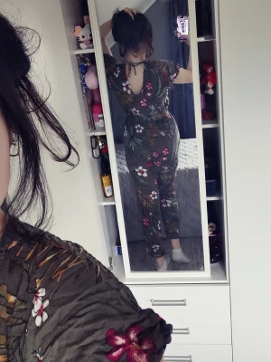 Grön blommig jumpsuit med v-ringning - Säljer en grön jumpsuit med tropiskt blommönster i rött, vitt och gult. Jumpsuiten har v-ringning fram och bak, korta ärmar, resår i midjan och ankellånga ben med resår vid benslut. Perfekt för dig som vill ha en chill och snygg look.