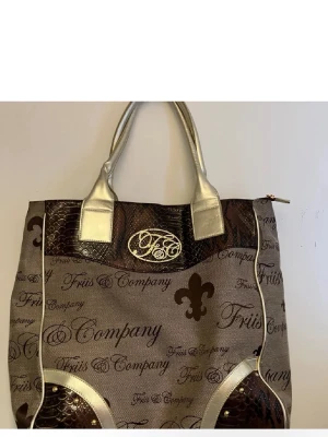 Friis & Company handväska - Stilren handväska från Friis & Company i beige tyg med bruna ormmönstrade detaljer och guldfärgade handtag. Väskan har tryckt logotyp och text, samt dekorativa mönster och guldfärgade detaljer. Perfekt för dig som gillar unika accessoarer.