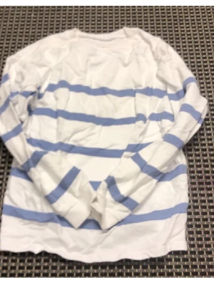 Vit tröja med blå ränder - Säljer en vit långärmad tröja med ljusblå horisontella ränder. Tröjan har rund halsringning och små slitsar i sidorna nedtill. Perfekt för en avslappnad och stilren look. Materialet känns mjukt och bekvämt mot huden.