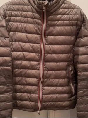 Moncler David down olivgrön - Lätt och stilren olivgrön dunjacka med horisontella sömmar och en snygg randig dragkedja framtill. Jackan har hög krage och två sidofickor med dragkedja. Perfekt för kalla vinterdagar och enkel att matcha med olika outfits.                                                    Skick 7/10 storlek 3