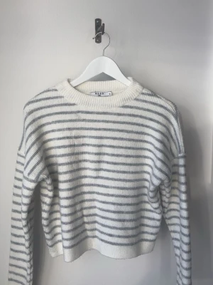 Randig stickad tröja från NA-KD - Säljer en vit stickad tröja med tunna grå ränder från NA-KD. Tröjan har en avslappnad passform, rund halsringning och långa ärmar. Perfekt för lager på lager och riktigt mysig till höst och vinter.