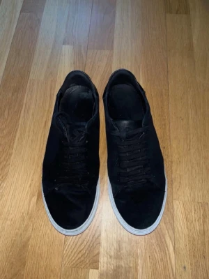 Svarta låga sneakers med vit sula - Snygga svarta sneakers med klassisk låg profil och vit sula. Skorna har svart ovandel i mocka eller liknande material och matchande svarta skosnören. Perfekta för en clean och stilren look.