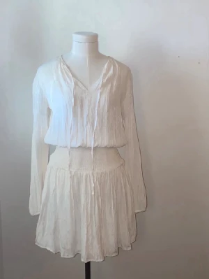 Vit omlottklänning med knyt - Från anine bings kollektion med Gina tricot. Exklusiv. Luftig vit klänning med omlott och knyt i halsen. Klänningen har långärmade ärmar, v-ringad hals och smockad midja för en bekväm passform. Kjolen är lätt utställd och materialet känns mjukt och svalt, perfekt för varma dagar.