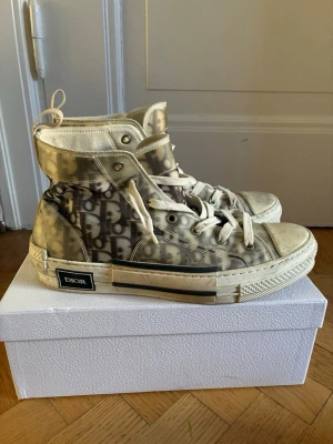 Beige Dior B23 high-top sneakers - Säljer ett par beige Dior B23 high-top sneakers med ikoniskt monogrammönster på sidan. Skorna har genomskinliga paneler, vita snören och robust sula med svart Dior-logga på hälen. Materialet är en mix av canvas och syntet. Perfekt för dig som vill sticka ut 