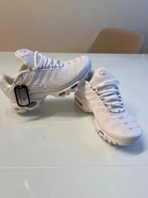 Nike Air Max Plus vita sneakers - Nike Air Max Plus sneakers i helvitt med mesh- och syntetmaterial. Skorna har den ikoniska vågiga designen, synlig Air Max-dämpning i sulan och vita snören. Perfekta för dig som gillar en clean och sportig look.