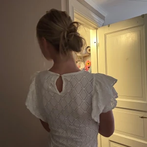 Vit topp med volangärmar och struktur - Snygg vit topp med volangärmar och ett diskret rutmönster i tyget. Toppen har en liten öppning bak i nacken. Perfekt till jeans eller kjol för en fräsch look och klassig lock. 