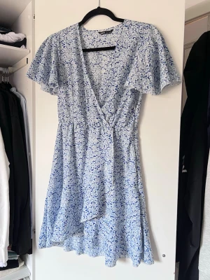 Blommig blå omlottklänning, stl S - Söt ljusblå omlottklänning med blommigt mönster i vitt och blått. Korta, fladdriga ärmar och resårband i midjan.