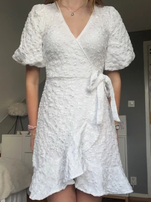 Söt vit omlottklänning - studentklänning - Supersnygg vit omlottklänning med puffärm och volang från Pieces som inte säljs i butik längre!🥰 Passar perfekt till studenten eller sommaren! Helt ny med prislapp kvar😍 Säljer då den aldrig kommit till användning😔