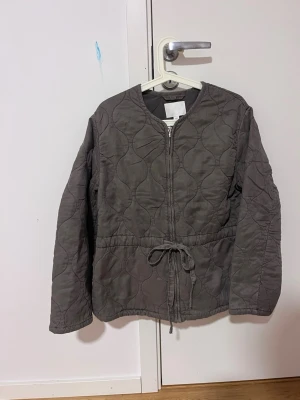 Populär Grå quiltad vår jacka med knyt - Quiltad grå cardiganjacka med rund halsringning och dragkedja framtill. Jackan har ett knytband i midjan för justerbar passform och ett diskret mönster i tyget. Perfekt för lager-på-lager och en chill look. Kan också ta ut snöret om man vill, original pris 649 kr ! Helt ny med prislapp!