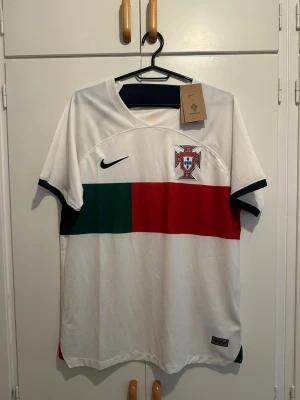 Portugal Nike fotbollströja vit - Säljer en vit Portugal fotbollströja från Nike med korta ärmar. Tröjan har gröna och röda blockdetaljer på framsidan, Portugals landslagsemblem på bröstet och svart Nike-logga. Tillverkad i lätt och ventilerande material, perfekt för träning eller match.