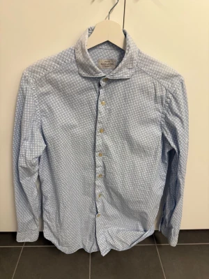 Ljusblå skjorta från Rose & Born, stl 39 - Snygg ljusblå skjorta från Rose & Born med små vita blommor som mönster. Skjortan har klassisk krage, knappar framtill och långa ärmar. Perfekt för dig som vill ha en stilren men ändå unik look.