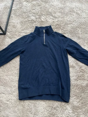 Mörkblå stickad tröja med halvzip - Mörkblå stickad tröja från Jack & Jones med halvzip och hög krage. Tröjan har långa ärmar och ribbade muddar vid ärmslut och nederkant. Perfekt för lager på lager och enkel att matcha med jeans eller chinos. OBS! Barnstorlek 152.
