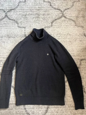 Mörkblå polotröja från Morris - Mörkblå stickad polotröja från Morris med ribbade muddar och hög krage. Tröjan har en liten broderad logga på bröstet och är tillverkad i mjukt material. Perfekt för lager på lager och stilrena outfits.