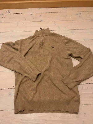 Beige halvzip tröja från Gant - Snygg beige stickad tröja från Gant med halvzip och liten broderad logga på bröstet. Tröjan har ribbade muddar vid ärmslut och nederkant samt hög krage. Perfekt för lager på lager och en clean stil.
