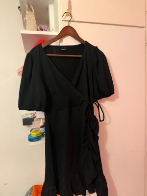 Svart omlottklänning Gina Tricot - Snygg svart omlottklänning från Gina Tricot med puffiga korta ärmar, knyt i midjan och volangkant nertill. Klänningen har en v-ringad hals och är tillverkad i ett mjukt, ribbat material som ger en skön känsla.